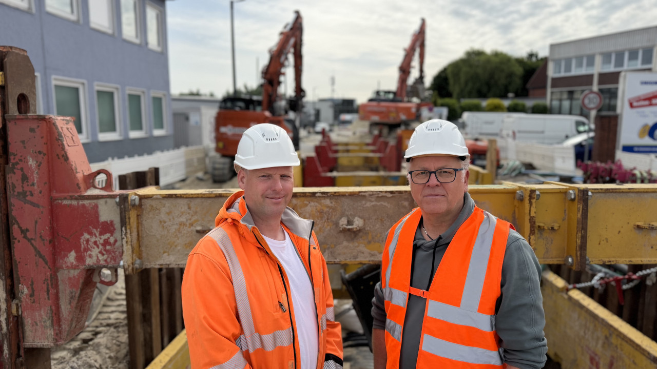 Zwei Personen in orangenen Warnwesten vor einer Baustelle.
