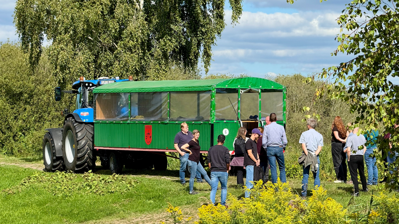 Personengruppe vor einem Traktor mit Planwagen, umgeben von grüner Natur.