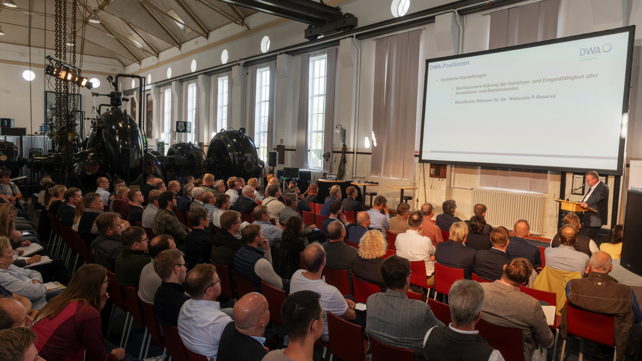 Eine Halle mit vielen Menschen, die auf Stühlen sitzen und zur Bühne schauen.