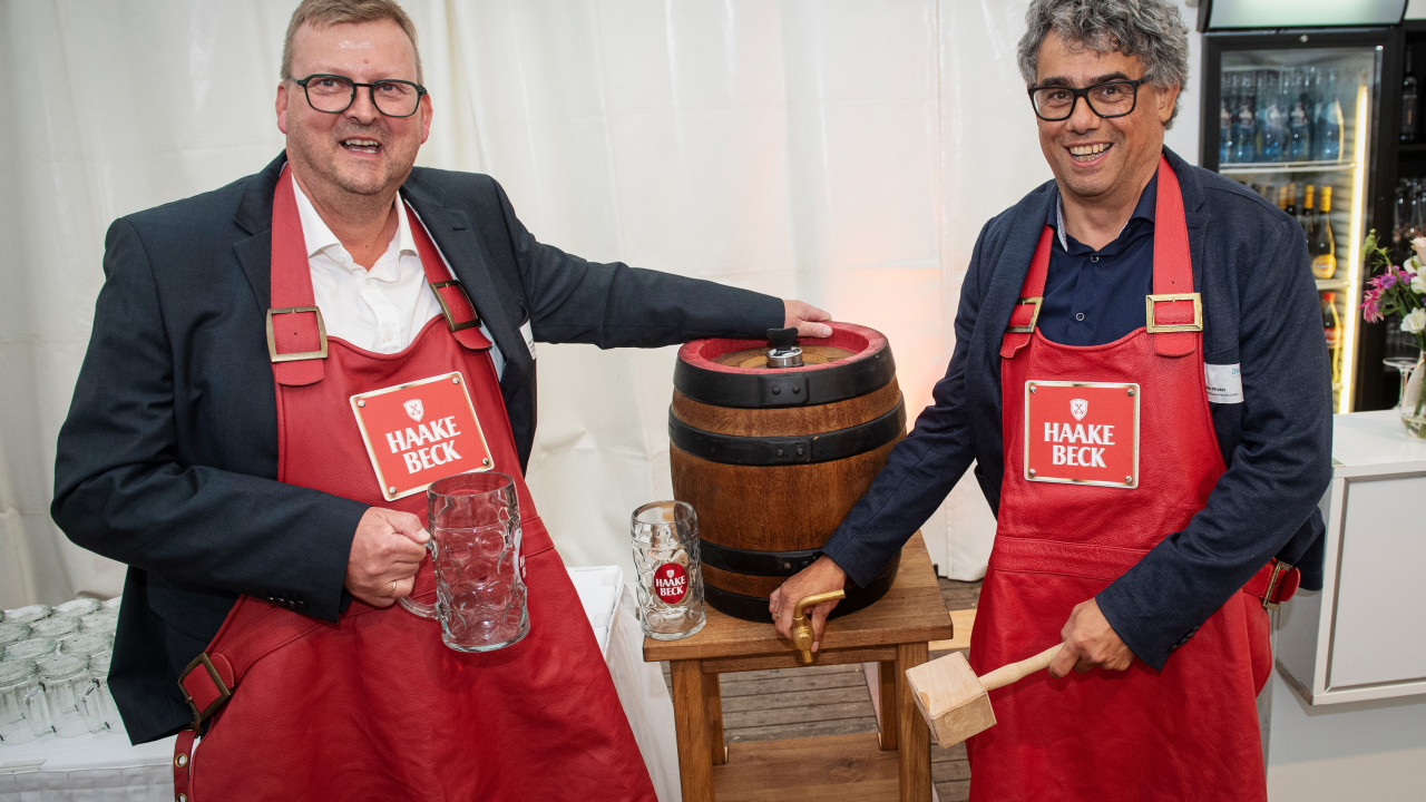 Zwei Männer stehen neben einem Bierfass mit Gläsern und einem Hammer in der Hand.