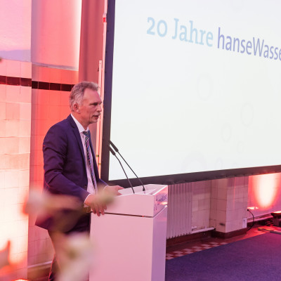 Der ehemalige Geschäftsführer Uwe Dahl auf einer rosa beleuchteten Bühne, hinter ihm ein Bildschirm mit "20 Jahre hanseWasser" darauf