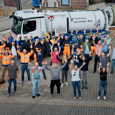 Szene aus dem Unternehmensfilm, eine große Gruppe von Mitarbeiter*innen reißt jubelnd die Arme empor, von leicht oben mit etwas Abstand vor einem Spülwagen auf dem Betriebshof in Findorff aufgenommen.