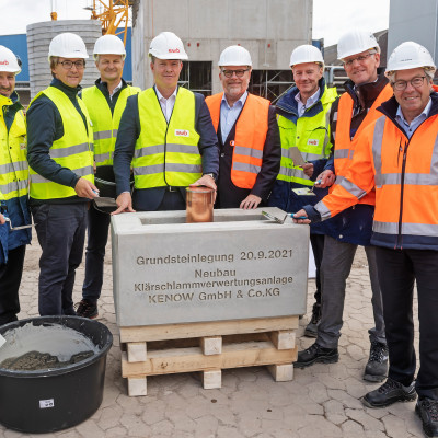 Mitarbeiter von swb und hanseWasser stehen um einen symbolischen Grundstein, auf dem das Datum 20.09.2021 und "Neubau Klärschlammverwertung KENOW" steht. SIe versenken einen kupferfarbenen Zylinder im offenen Stein..